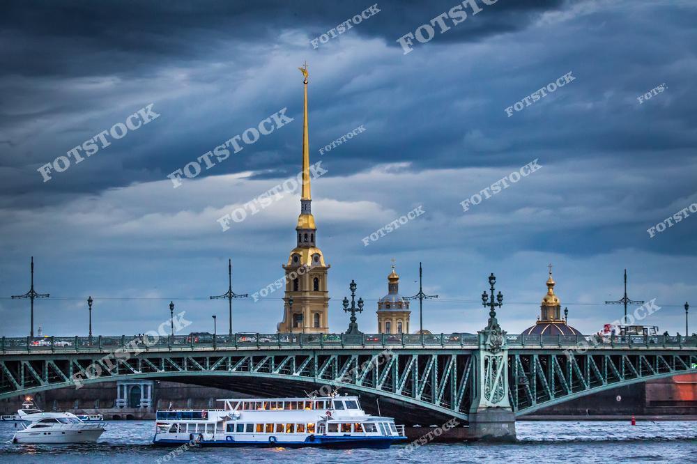 На фотографии изображен Троицкий мост, соединяющий берега Невы, а за ним возвышается золотой шпиль Петропавловского собора. По реке плывут туристические катера. Этот пейзаж отражает неповторимую атмосферу петербургской погоды и классические виды города.